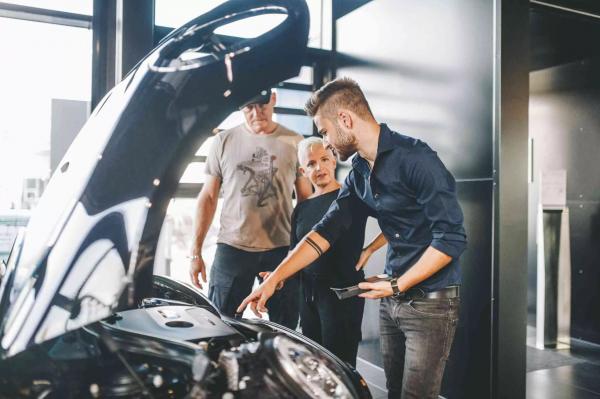 Ein Kundenberater einer MINI Servicewerkstatt erklärt einem älteren Ehepaar einen Sachverhalt an ihrem MINI bei offener Motorhaube.