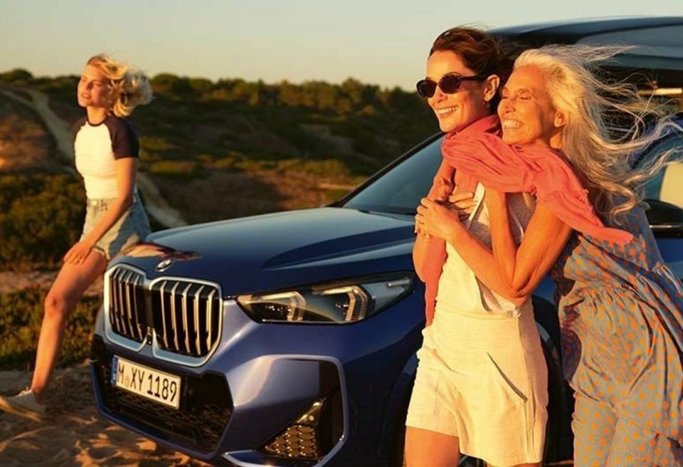 Tochter, Mutter und Großmutter laufen in Richtung Abendsonne an ihrem BMW X1 in "phytonicblau metallic" vorbei.