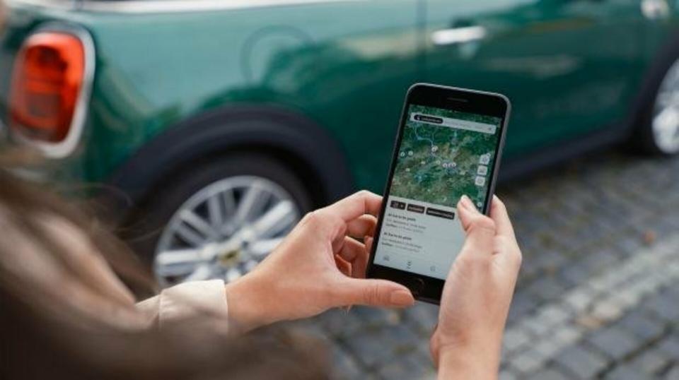 Ein Frau betrachtet in der Hand ihr Smartphone mit der MINI App. Im Hintergrund steht sein MINI Cooper.