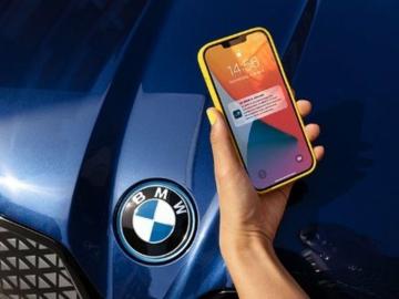 Eine weibliche Hand mit gelb lackierten Fingernägeln hält ein gelbes Smartphone vor die Motorhaube ihres BMW mit der My BMW App.