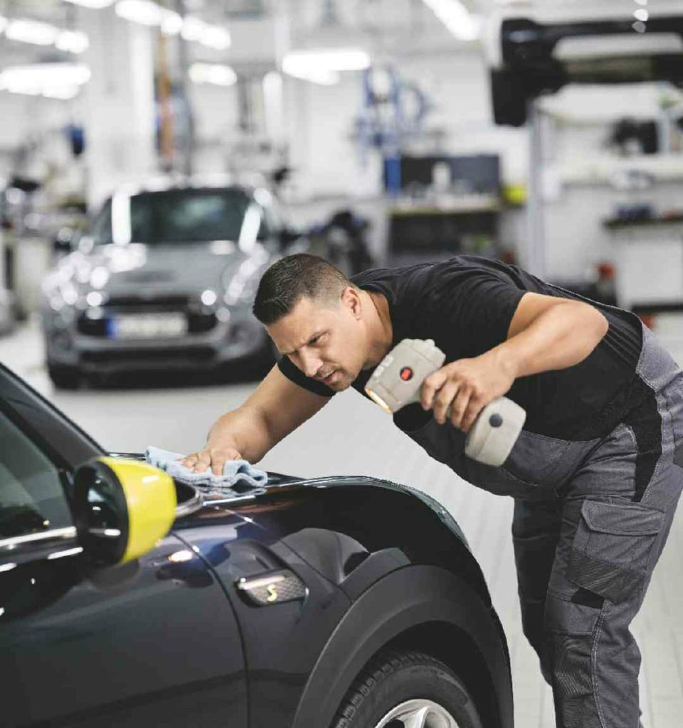 Ein Mitarbeiter eine MINI Servicewerkstatt prüft den Lack der Motorhaube eines schwarzen MINIs mit einer Lampe.
