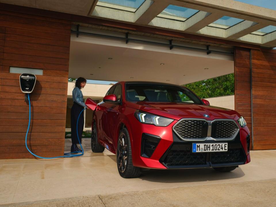Ein BMW iX2 in "fire red metallic" wird in einer Garageneinfahrt von einer jungen Dame an einer Wallbox aufgeladen.