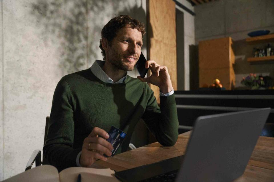 Ein junger Mann sitzt an seinem Schreibtisch vor seinem Computer, hält eine Kreditkarte in der Hand und telefoniert.