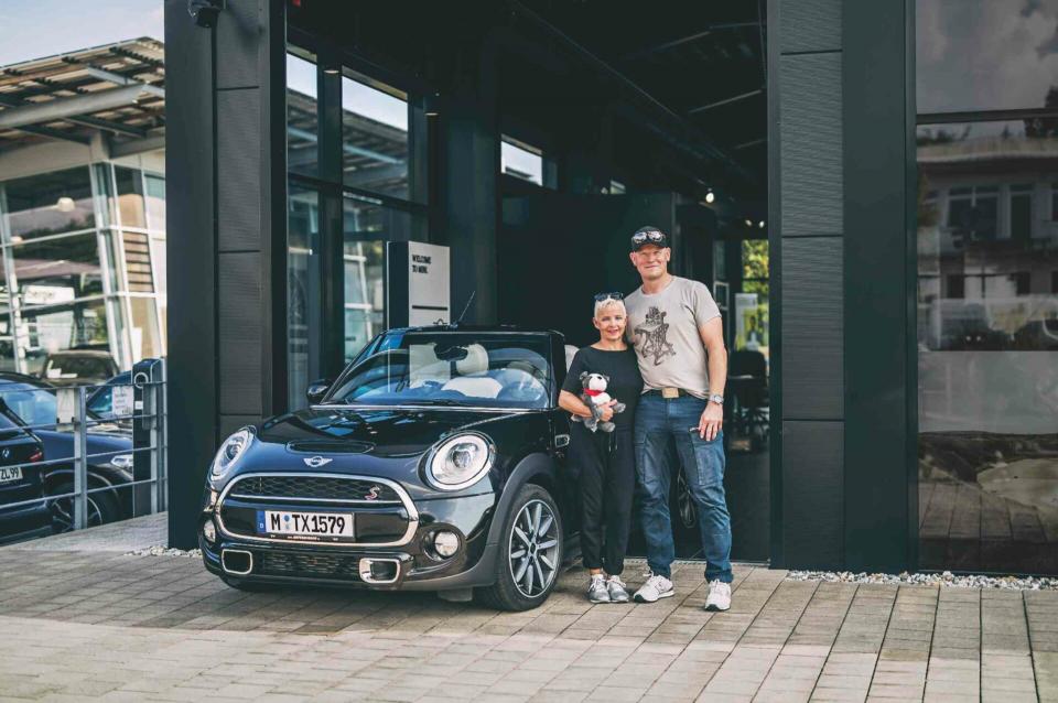 Ein älteres Ehepaar steht glücklich mit einem Tedddybären in der Hand vor ihrem MINI Cabrio an der Service-Werkstatt.
