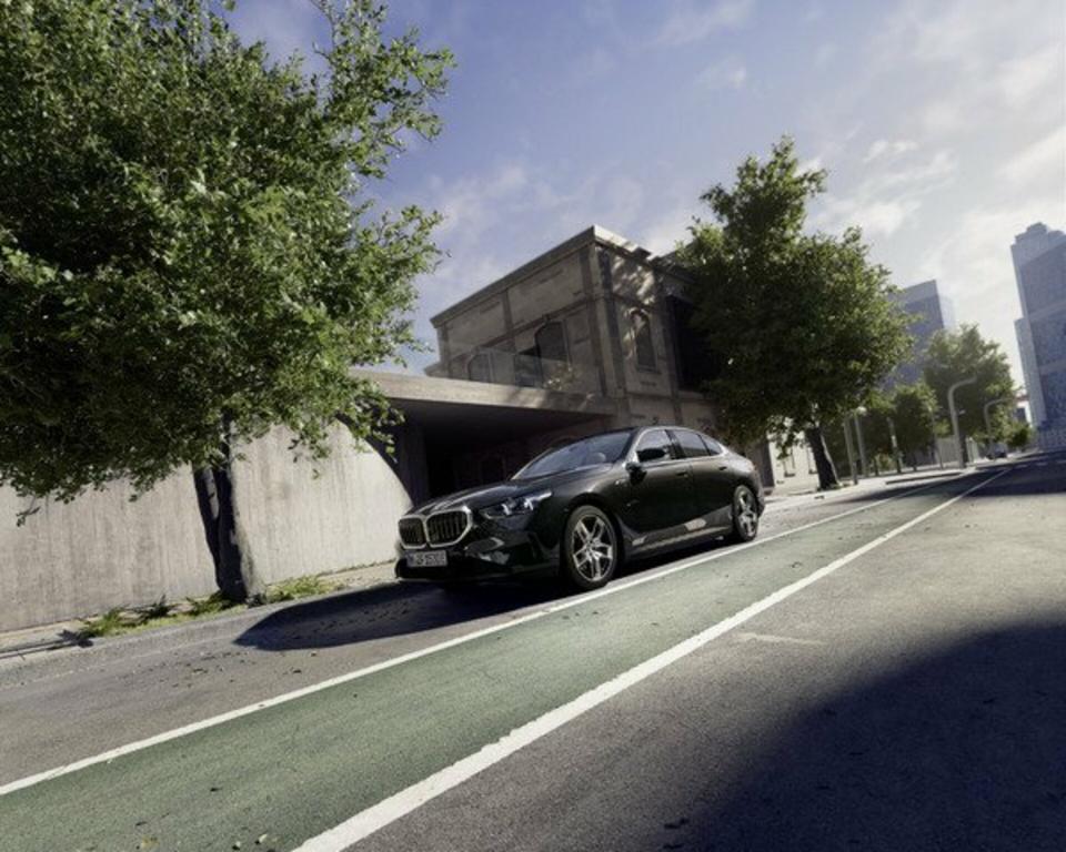 Ein schwarzer BMW 5er parkt auf dem Parkstreifen neben einem älteren Industriegebäude. An der Straße stehen mehrere grüne Bäume.