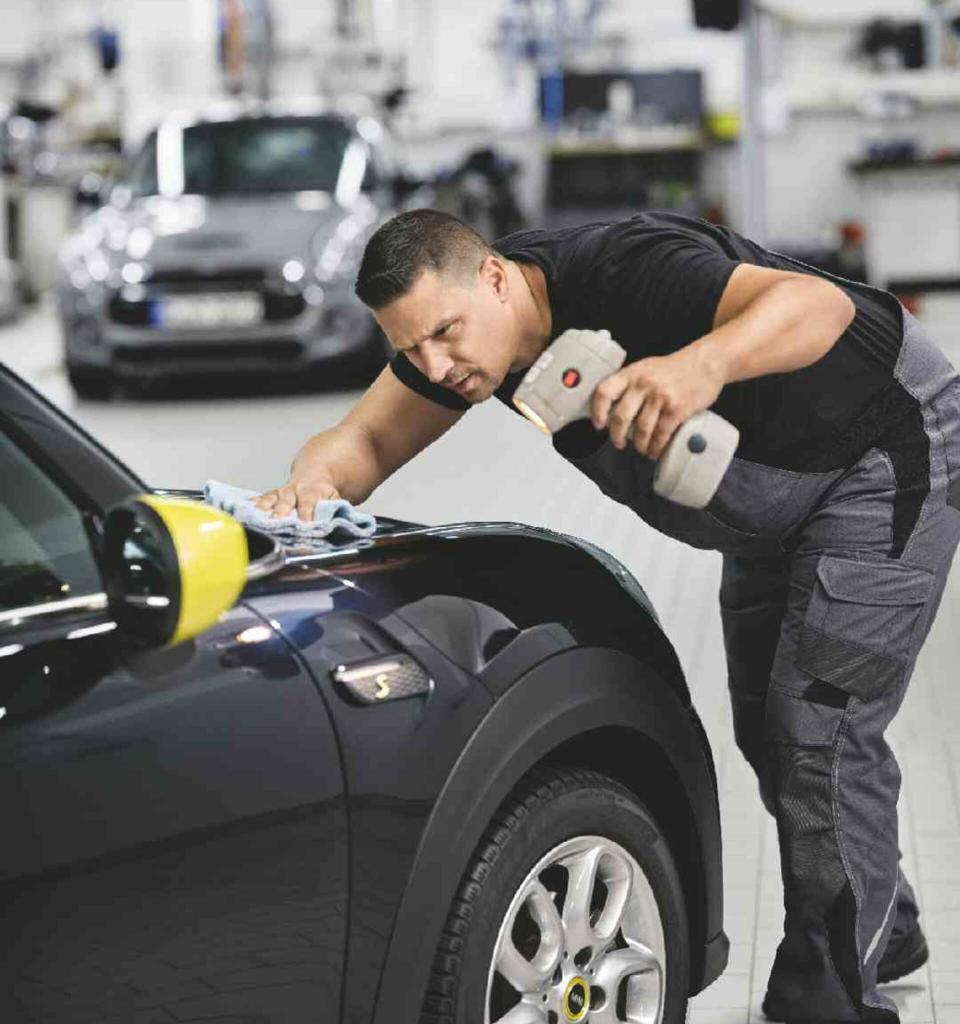 Ein Mitarbeiter eine MINI Servicewerkstatt prüft den Lack der Motorhaube eines schwarzen MINIs mit einer Lampe.