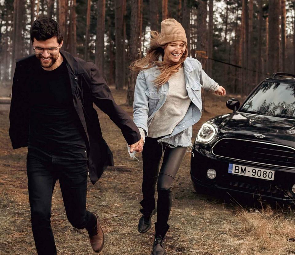 Ein glückliches Pärchen läuft händchenhaltend vor ihrem schwarzen MINI Cooper durch den Wald.
