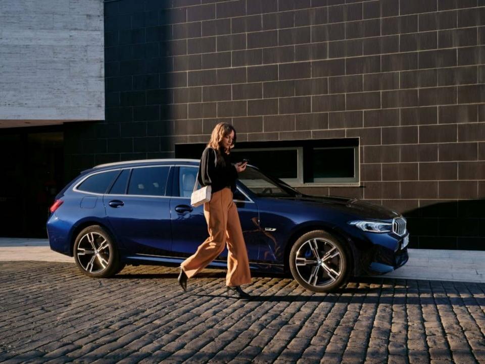 Eine Businessfrau läuft an ihrem BMW 3er in "transitblau metallic" vorbei und sieht auf ihr Smartphone.