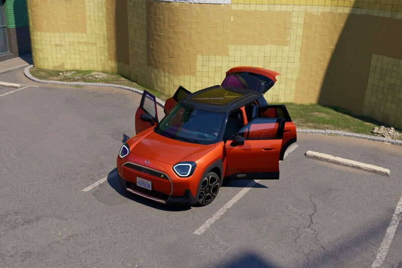 Ein MINI Aceman Electric in der Farbe Rebel Red parkt mit allen Türen geöffnet vor einem wellenförmigen Gebäude auf dem Parkplatz.