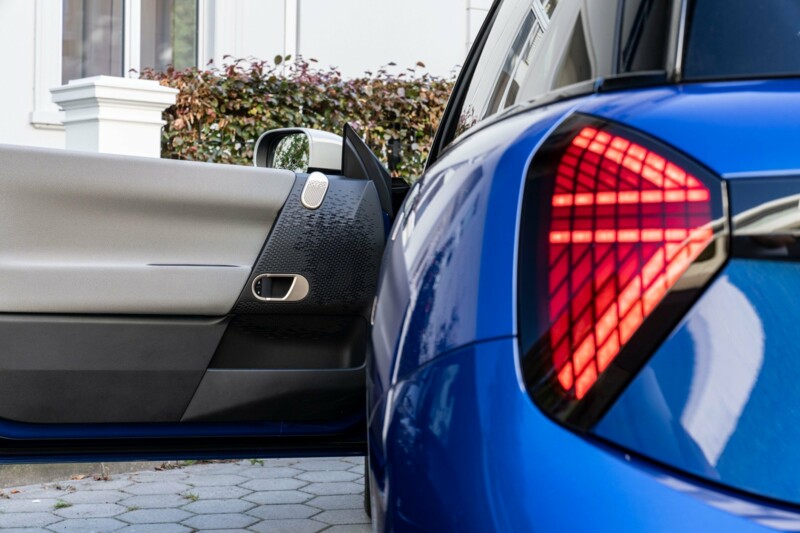 Ein MINI Cooper Electric in der Farbe blazing blue mit geöffneter Fahrertür parkt neben einem Wohnhaus.