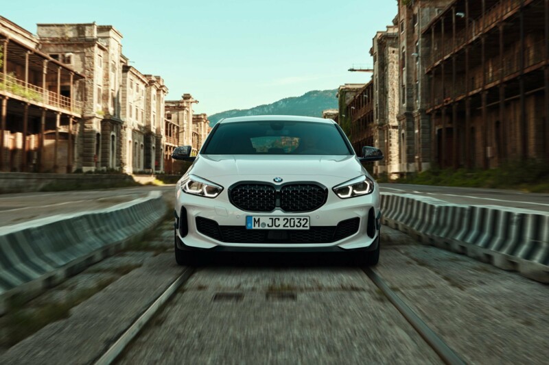 Ein weißer BMW 1er fährt in industriell-urbaner Umgebung auf stillgelegten Schienen.