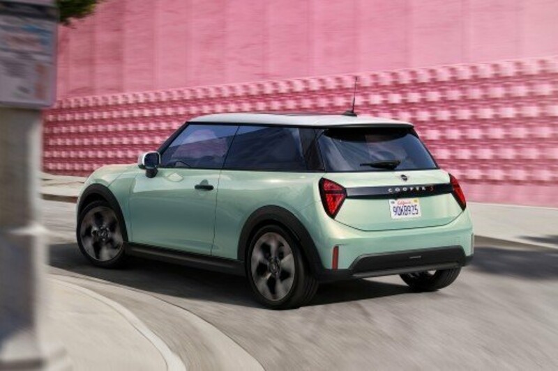 Ein MINI Cooper 3-Türer in der Farbe Ocean Wave grün fährt neben einem pinkfarbenen Gebäude um die Kurve.