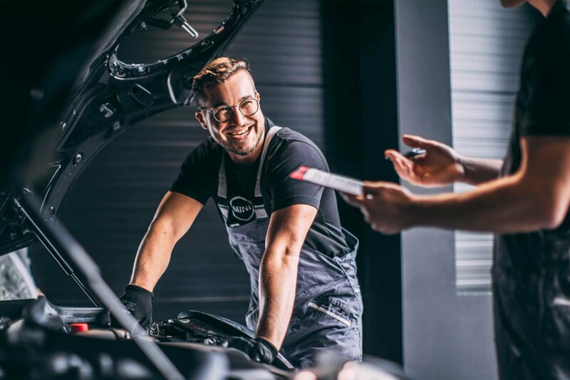Zwei Mitarbeiter einer MINI Werkstatt haben Spaß während der Reparatur bei offener Motorhaube.
