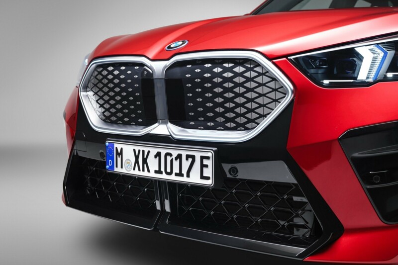 Die Frontansicht des neuen BMW iX2 in der Farbe Fire Red metallic vor einer Fotokulisse.