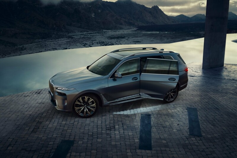 Ein BMW X7 xDrive40i mit geöffneter linken Hintertür an einem Kopfstein-gepflasterten Platz an einem Bergsee.