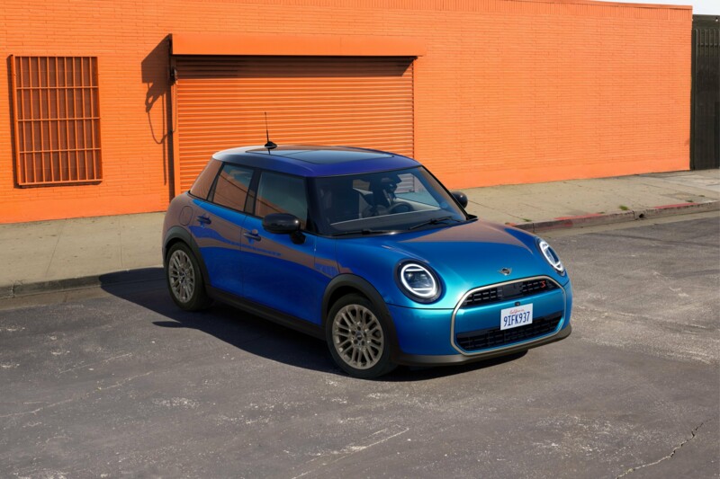 Ein MINI Cooper 5-Türer in der Farbe Blazing blue parkt and der orangefarbenen Fassade eines Lagergebäudes.